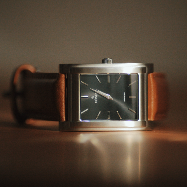 A close-up of a sleek rectangular watch with a dark face and silver frame. Featuring a brown leather strap, its minimalist design showcases clear hour and minute markers. The softly blurred background highlights this elegant timepiece.