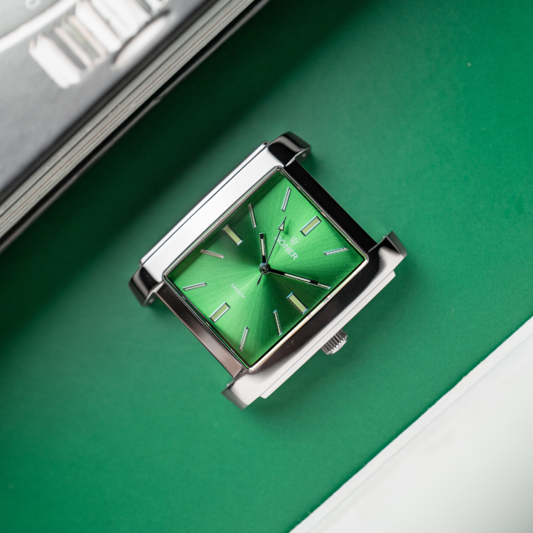 A stunning rectangular wristwatch with a vibrant green dial and silver indices, encased in stainless steel. Elegantly laid on a matching green surface beside the edge of a magazine, it exudes style and sophistication.