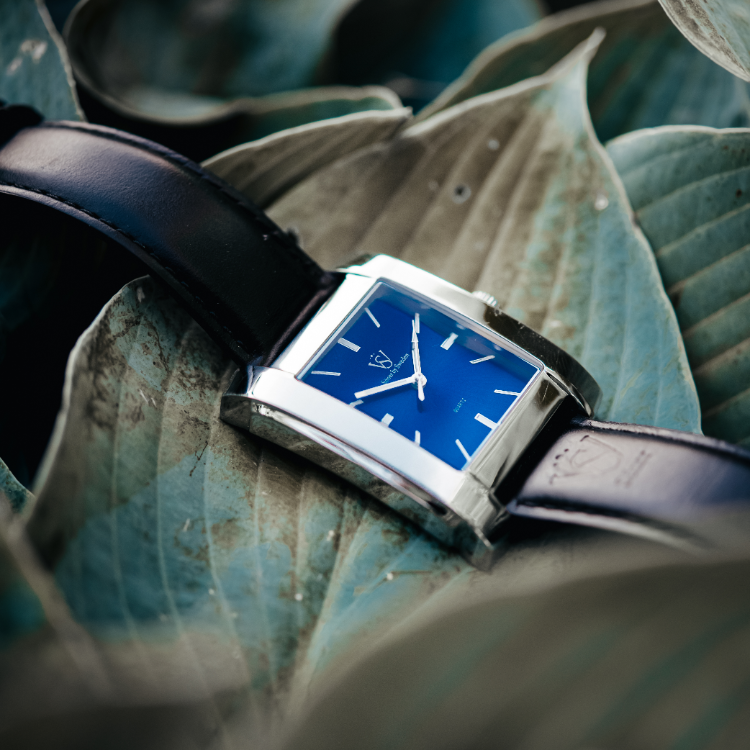 A sleek rectangular watch with a blue face and silver casing rests atop large, overlapping leaves. Featuring minimalist hour markers and a black leather strap, it contrasts elegantly with the cool, muted green leaves, their veins subtly enhancing the natural backdrop.
