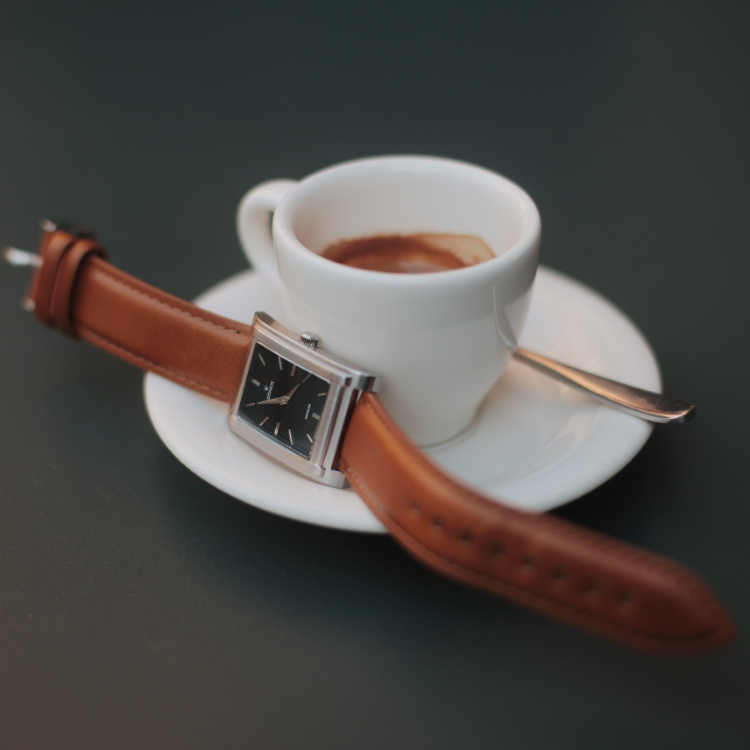 A rectangular watch with a brown leather strap encircles a white espresso cup on a saucer. A small spoon rests beside it, while the cup holds a partially drunk espresso against the dark surface background.