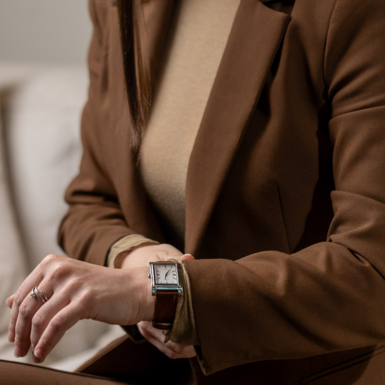 A person in a brown suit sits on a couch, checking the time on a rectangular watch with a brown leather strap. Their hand rests on their knee, and a beige turtleneck peeks out from under the suit jacket.