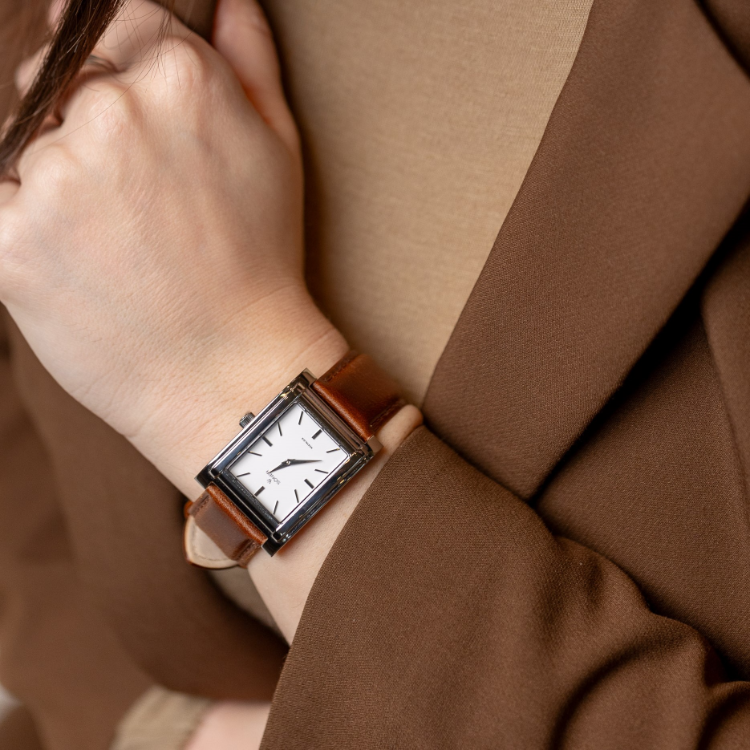 A person wearing a brown jacket and beige top is holding their arm. They are wearing a square-faced watch with a white dial and brown leather strap. The hand obscures part of the face.