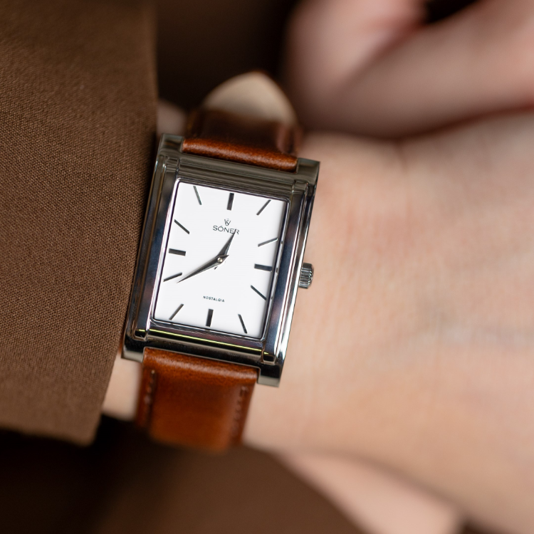 A close-up of a persons wrist wearing a rectangular, silver-framed wristwatch with a white dial, black hour markers, and a brown leather strap. The watch hands point to just before 9 oclock. The person is dressed in a brown sleeve.