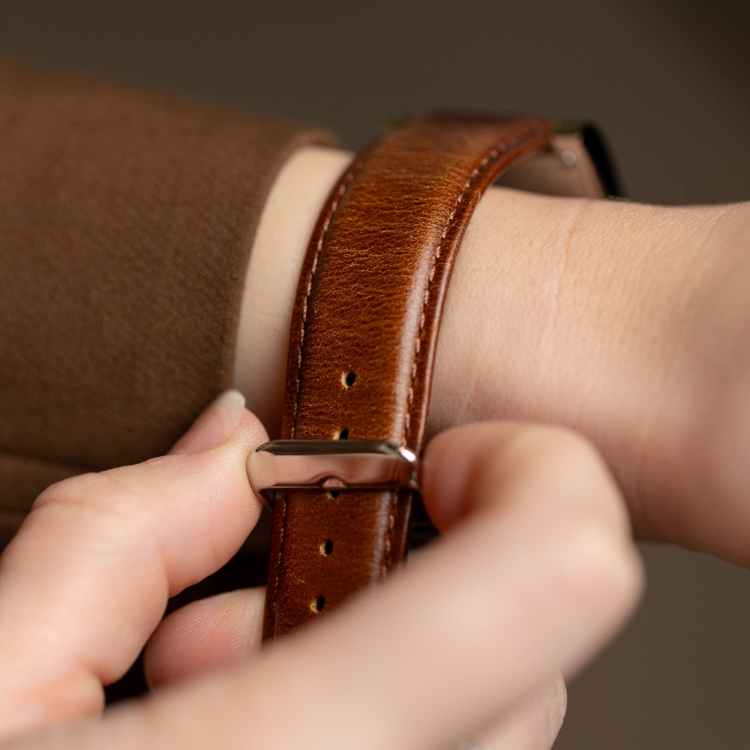 Close-up of a person adjusting a brown leather watch strap on their wrist. The watch has a polished silver buckle. The persons fingers hold the strap, and they are wearing a brown sleeve.