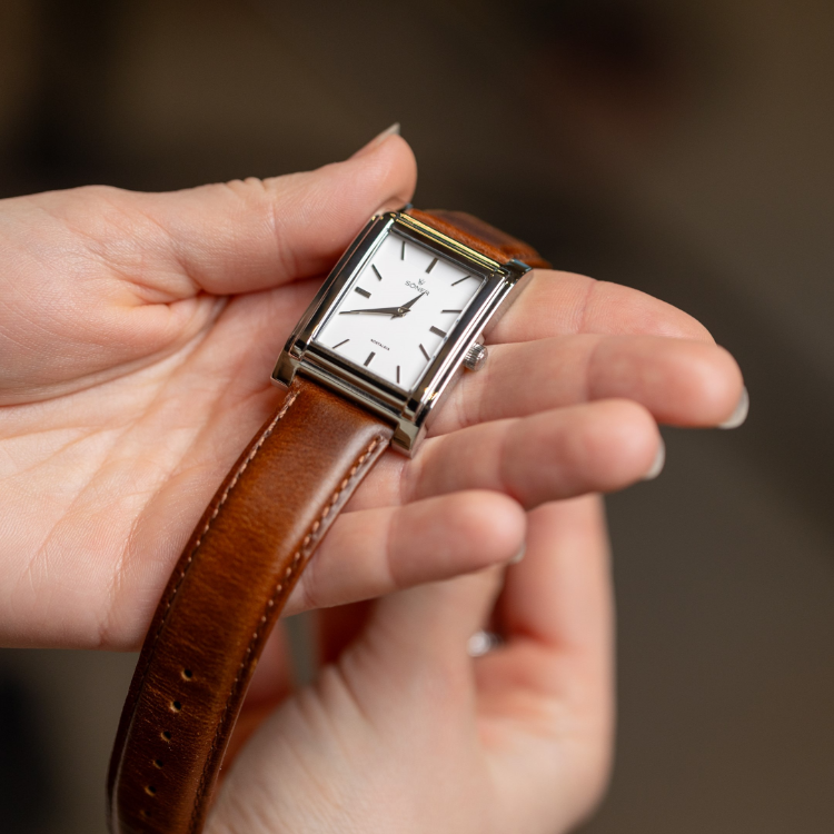 A person holds a rectangular wristwatch with a white face, black hour and minute hands, and a brown leather strap. The watch has a minimalist design with no numbers on the dial. The background is blurred.