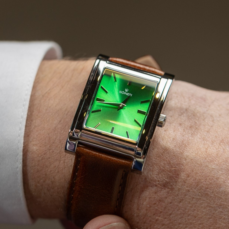 A close-up of a wrist adorned with a sleek rectangular watch. The vibrant green dial is encased in silver, paired with a brown leather strap. Its minimalist design features black hour markers and hands, while the word SONERI elegantly sits below the 12 oclock position.