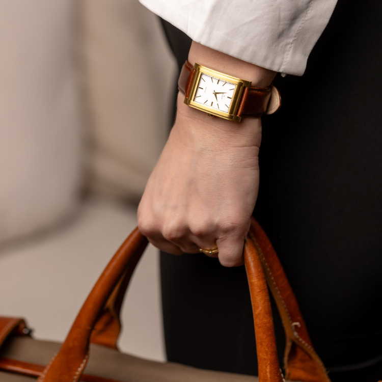 A person wearing a rectangular gold watch with a brown leather strap is holding a tan leather bag. Dressed in a white shirt and black pants, the subtle background hints at an indoor setting.