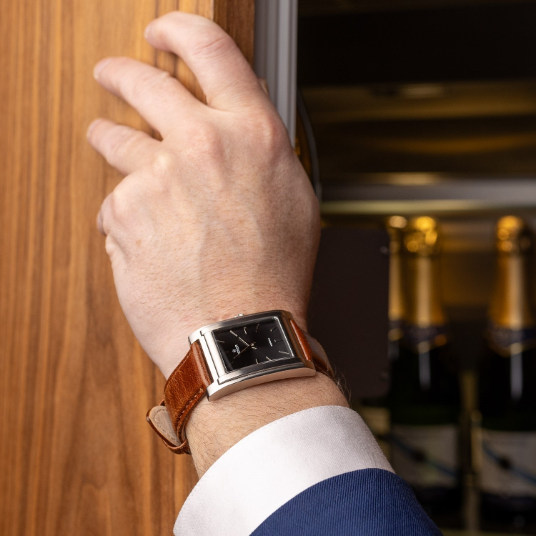 A person wearing a rectangular watch with a brown leather strap opens a wooden cabinet, revealing several bottles of champagne inside.