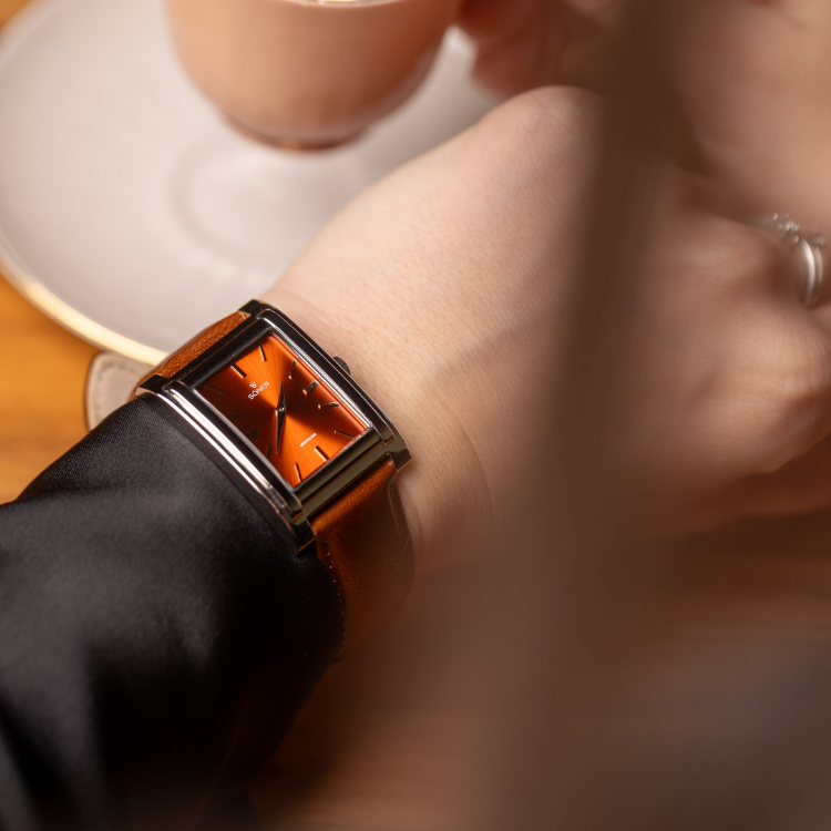 A person wears a sleek rectangular watch with an orange face and a brown leather strap, holding a cup on a saucer with a wooden table in the background. The minimalist design of the watch is in sharp focus.