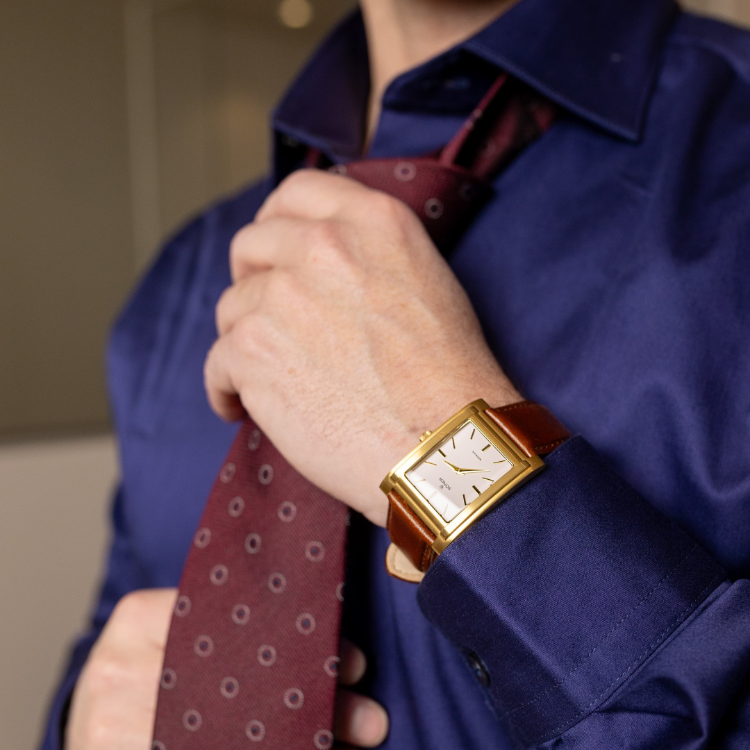 A person wearing a blue dress shirt adjusts a red patterned tie. On their left wrist, they sport a gold rectangular watch with a brown leather strap. The background is out of focus.