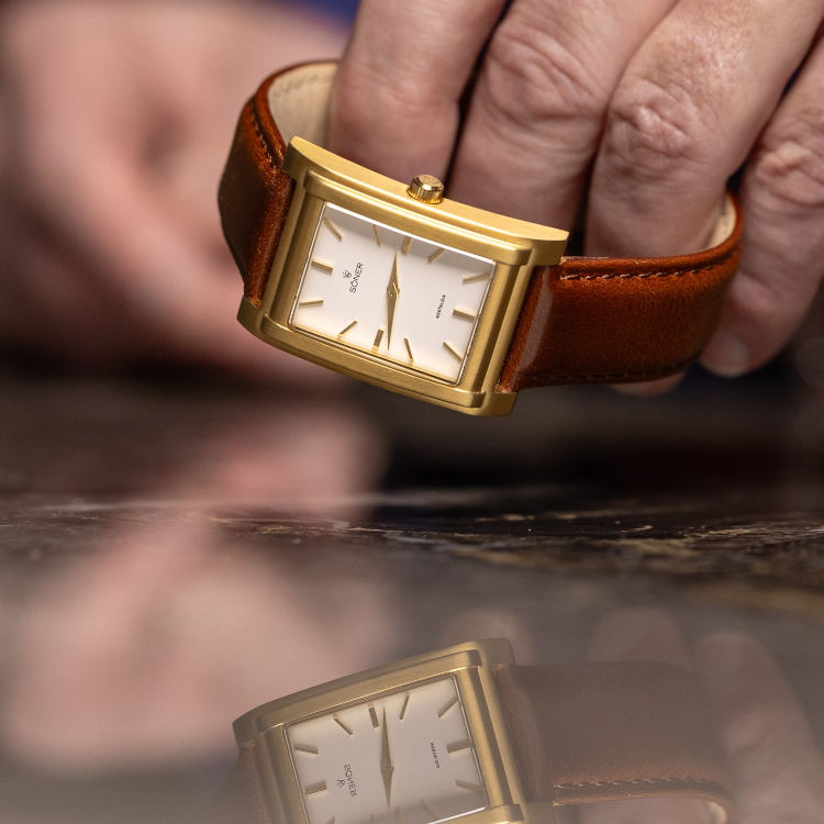 A close-up of a person holding a rectangular watch with a gold finish and brown leather strap. The cream face features simple gold hour markers and hands. Both the watch and hand are elegantly reflected on the shiny surface below.