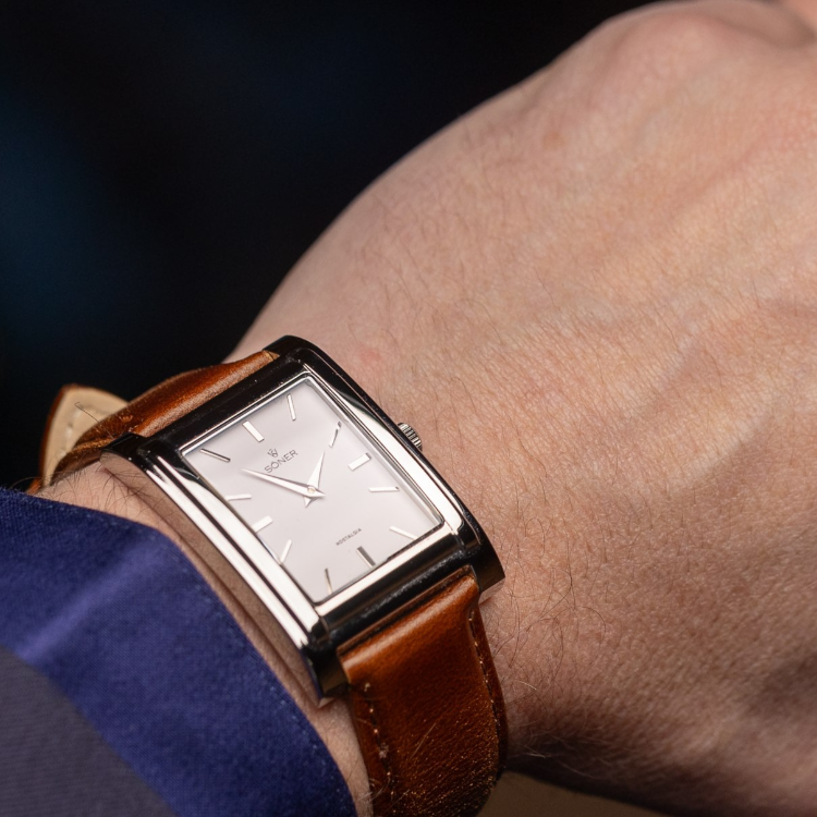 Close-up of a persons wrist wearing a rectangular watch with a silver frame and minimalist design. The white face features silver hour markers and hands, complemented by a brown leather strap. Paired with a blue shirt and brown jacket, its the perfect blend of style and sophistication.