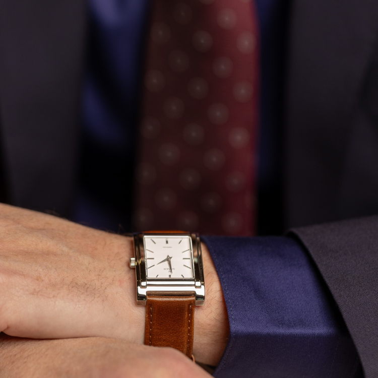 A close-up of a person wearing a suit with a blue shirt and a maroon polka dot tie. They are checking the time on a rectangular watch with a brown leather strap and a white dial.