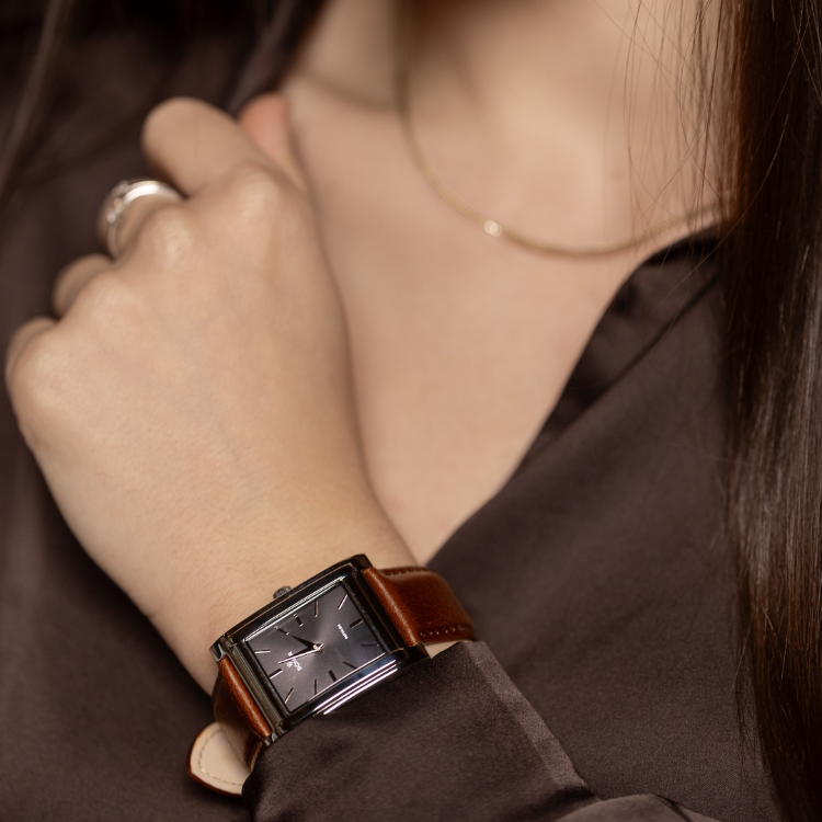Close-up of a person wearing a minimalistic rectangular watch with a brown leather band, set against a dark brown shirt. The hand, adorned with a gold chain and silver ring on the index finger, elegantly rests near the neck.