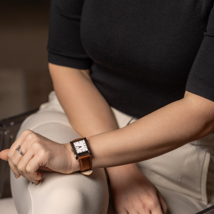 A person in a black top and beige pants is seated, with their rectangular watch featuring a brown leather strap glinting on their wrist as they lean on their knee. A ring adorns their left hand, all against a softly blurred background.