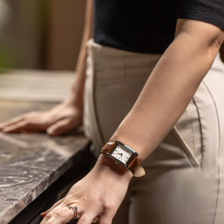 A person in beige pants and a black top leans on a marble surface. They wear a rectangular watch with a brown leather strap on their left wrist, and their right hand rests on their thigh.