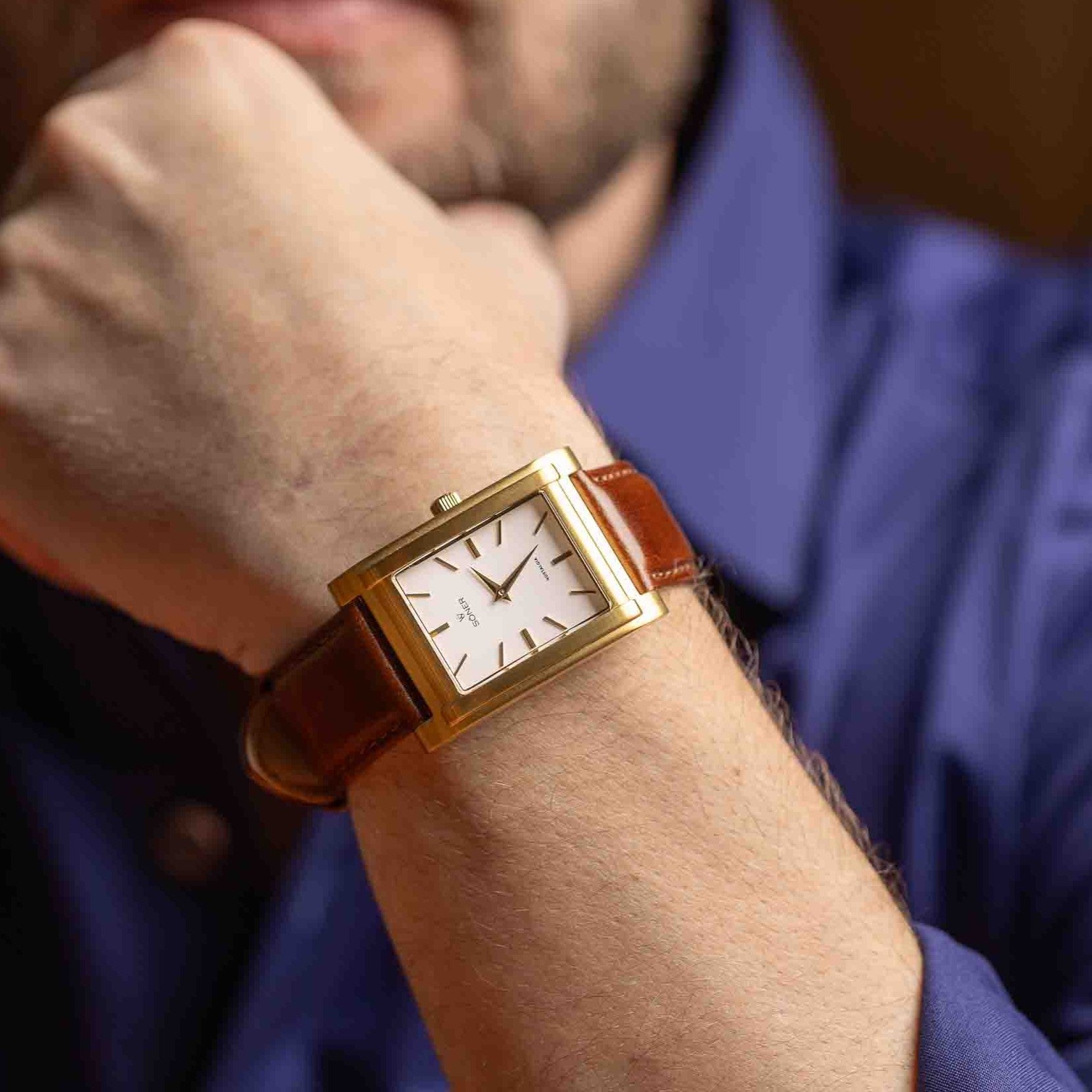 A person in a blue long-sleeve shirt rests their hand near their chin, wearing the NOSTALGIA NICE 11-Year Battery—a gold rectangular wristwatch with a brown leather strap and a white dial.