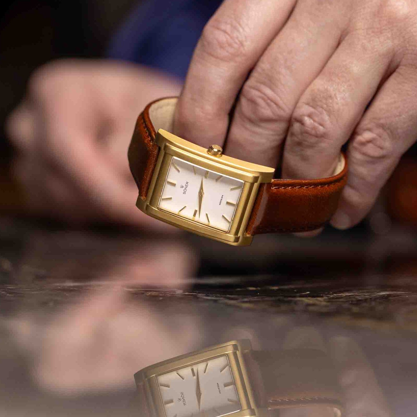 A hand holds the NOSTALGIA NICE wristwatch with a gold rectangular case, brown leather strap, and white face with gold hour markers and hands, above a reflective surface. Features an 11-year battery.