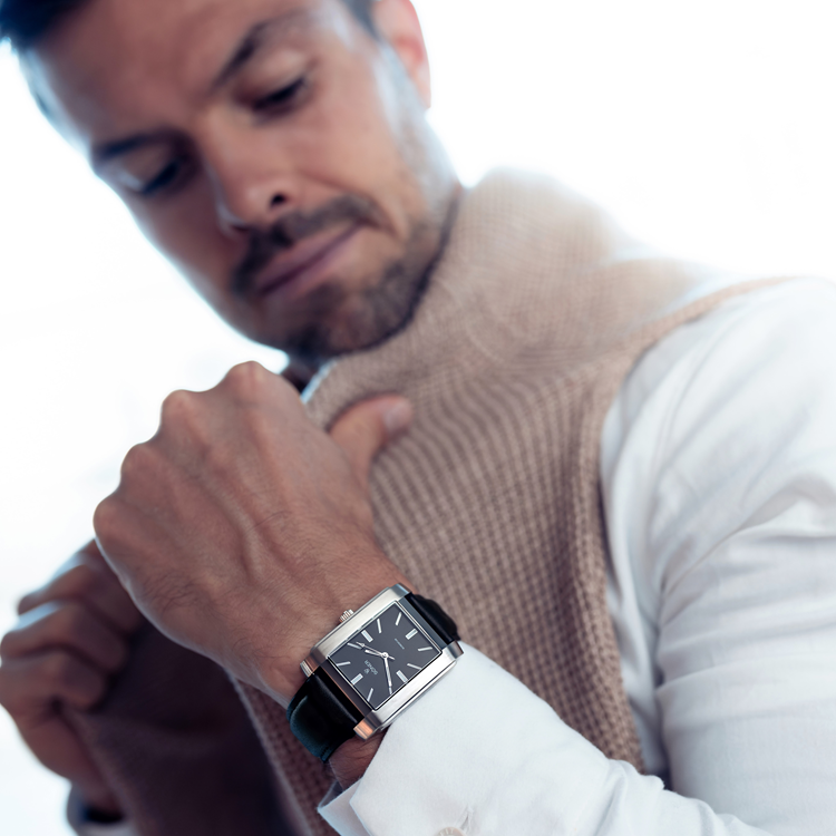 Man in a white shirt adjusts a beige sweater draped over his shoulders while looking down at his wristwatch. The watch has a square black face and a black strap. The background is softly blurred.