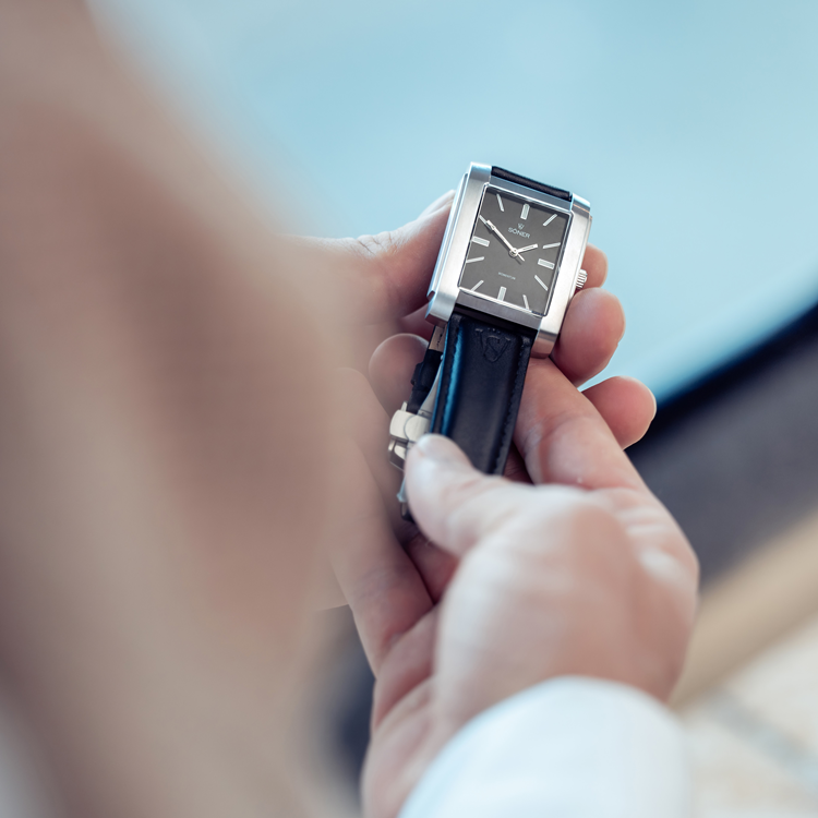 A person holding a rectangular, silver-framed wristwatch with a black leather strap. The watch has a black face with silver hour markers and hands. The background is blurred, focusing on the watch.