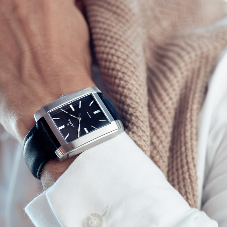 A person wearing a white shirt and a beige textured scarf showcases a sleek, rectangular watch. The black watch features a silver frame, black leather strap, minimalist design, and white hour markers.