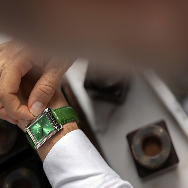 A close-up of a person adjusting a silver rectangular watch with a green square face and matching leather strap on their wrist. The individual is wearing a long-sleeve white shirt, set against a blurred background with indistinct objects visible.