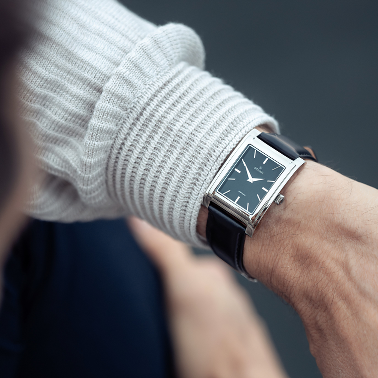 Close-up of a person wearing a rectangular black-faced wristwatch with a black leather strap. The person is wearing a light gray ribbed sweater, and the focus is on the watch. The background is blurred.