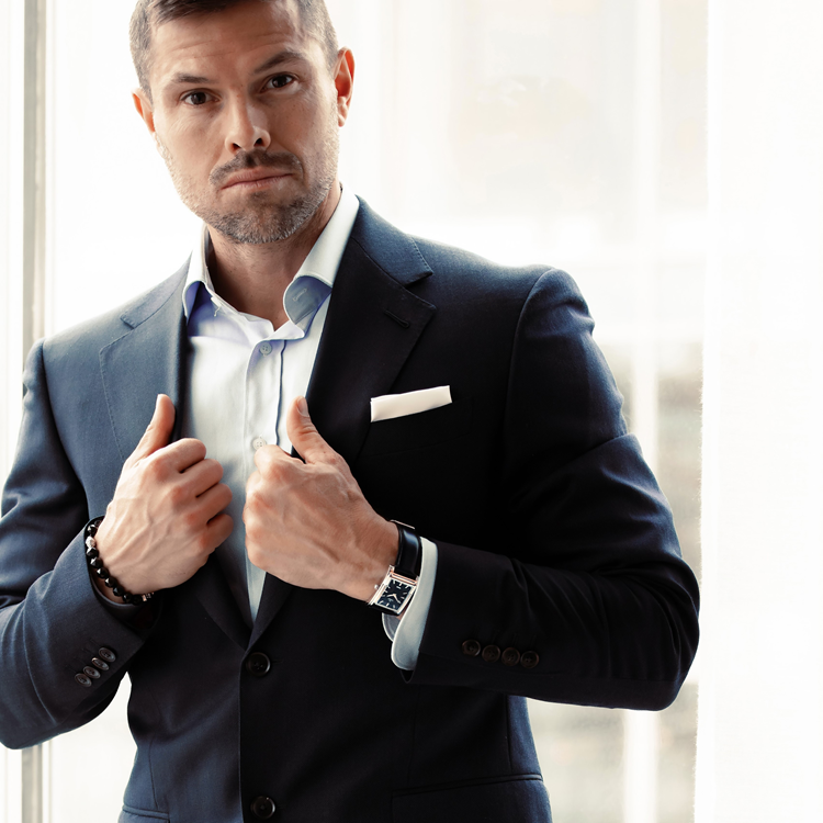 A man in a dark suit and white shirt stands by a sunlit window, adjusting his jacket. He wears a wristwatch and a beaded bracelet. A white pocket square is tucked into his breast pocket.