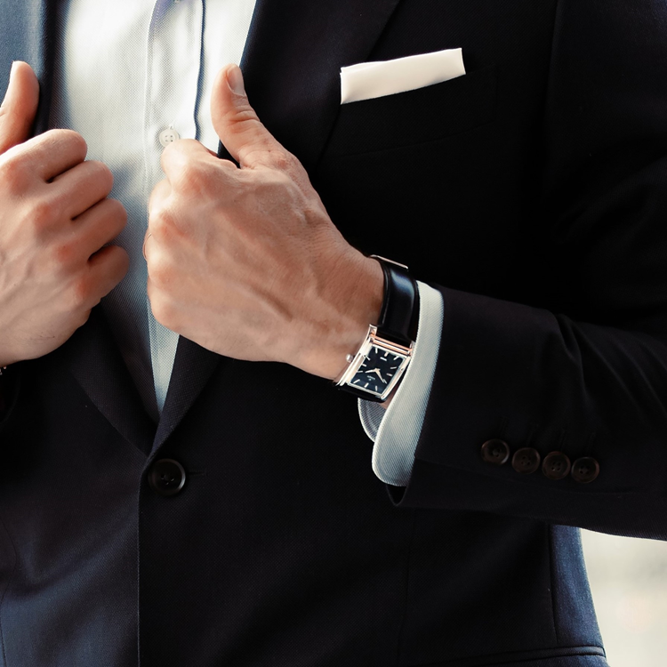 A person adjusts the lapels of a dark suit while wearing a light dress shirt. A sleek watch with a black leather strap is visible on their wrist, and a white pocket square is tucked into the jacket.