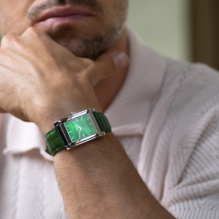 A person wearing a light-colored shirt sits with their hand resting against their chin, showcasing a stylish rectangular watch. The watch features a green square face and matching green strap on their wrist.