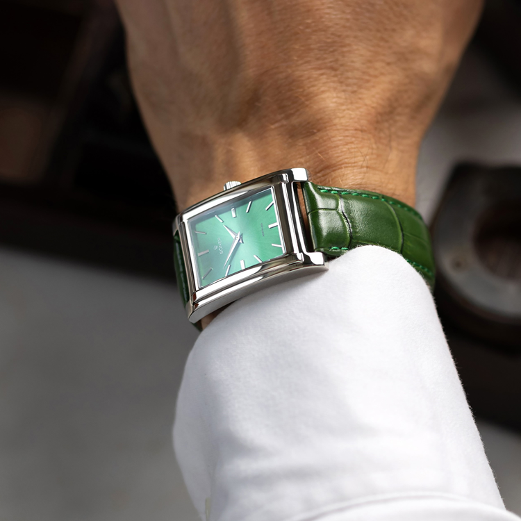Close-up of a person wearing a rectangular, green-faced wristwatch with a shiny metal frame and a green leather strap. The watch is worn over a white shirt sleeve.