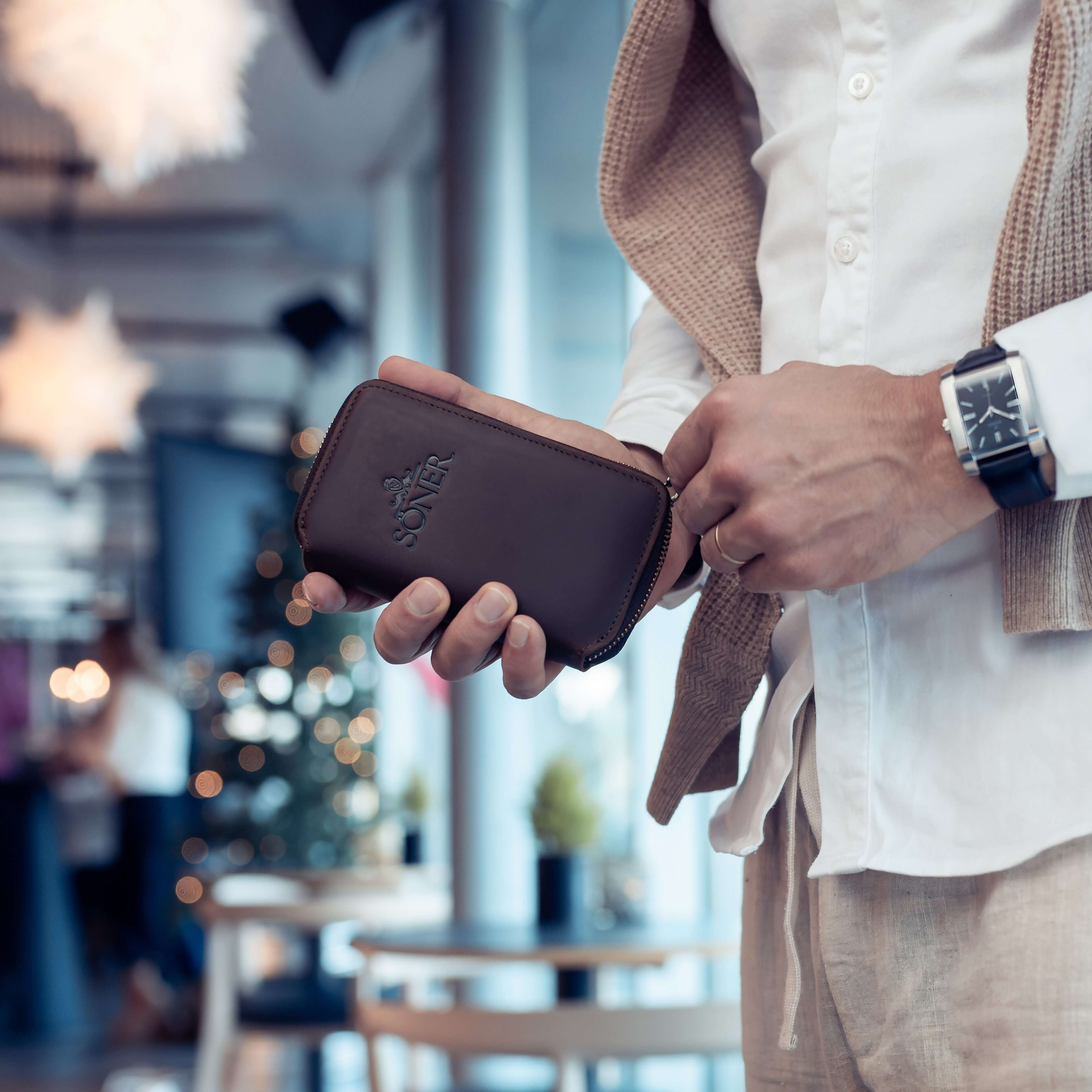 A person in a white shirt and beige pants holds a brown leather wallet. On their wrist is a watch protected by a Leather Watch Case. The background reveals a blurred interior with hanging pendant lights and subtle festive decor.