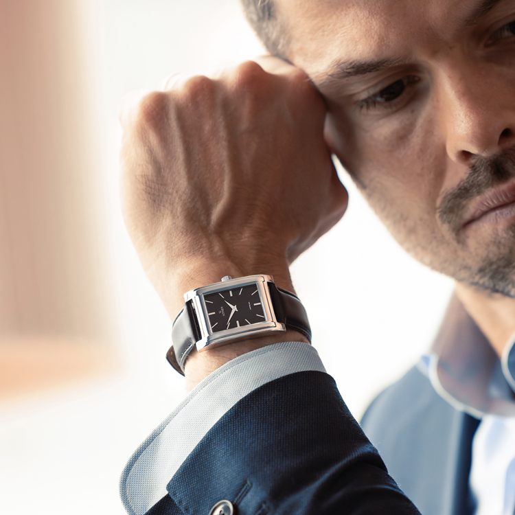 A close-up reveals a person wearing a sleek rectangular watch, its black and silver design complementing the leather strap. Dressed in a blue suit jacket over a white shirt, they gracefully rest their chin on their hand, gazing downward with an air of sophistication.