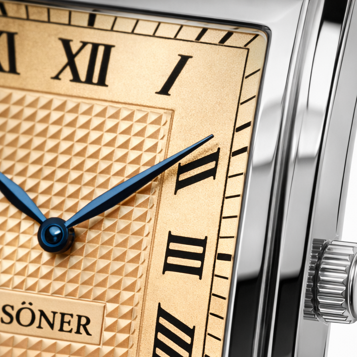 Close-up of a luxury watch face with a gold textured dial, black Roman numerals, and blue hands. The polished metal bezel and crown are partially visible on the right side.