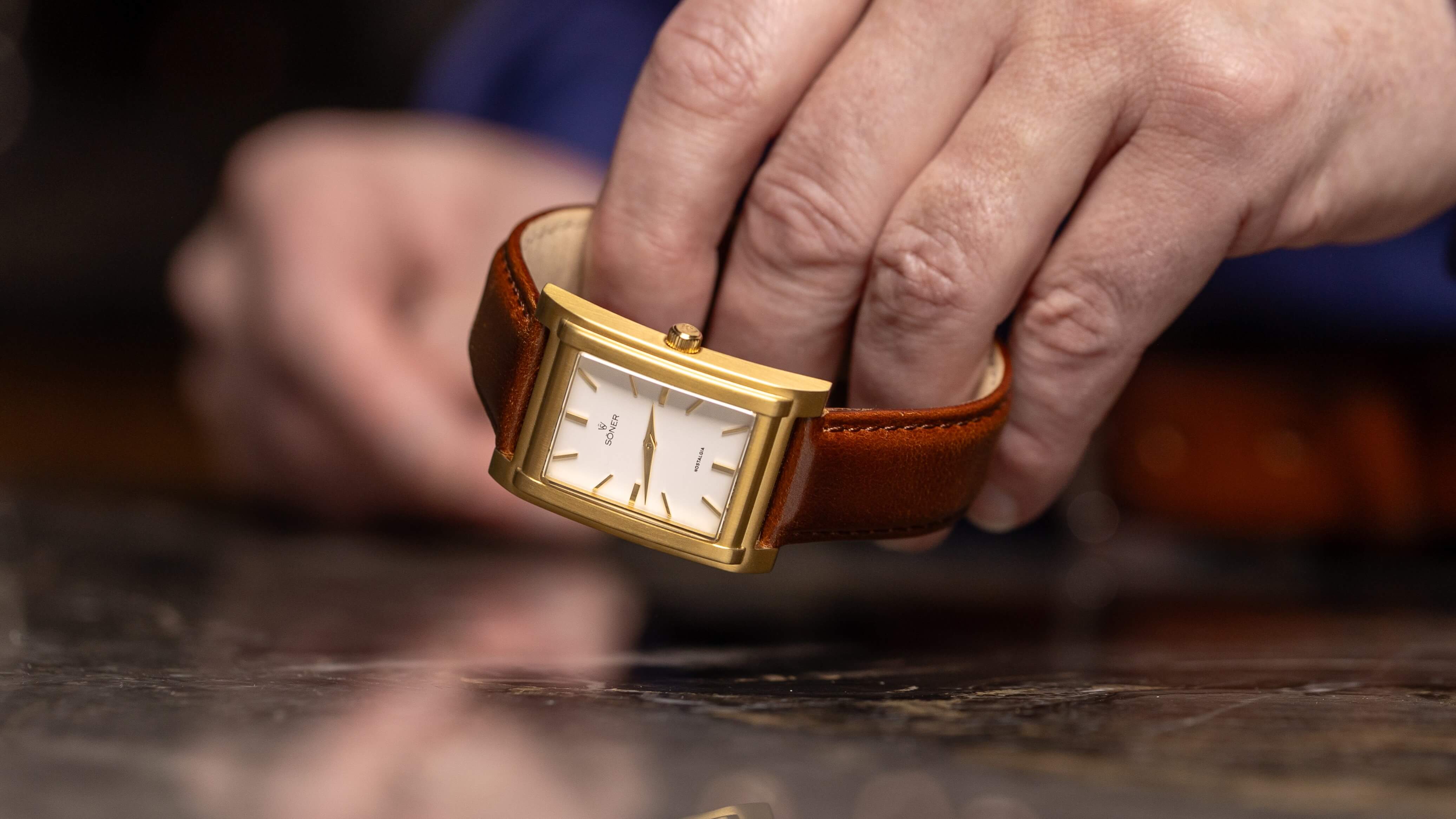 A person holding a rectangular gold wristwatch with a brown leather strap admires its reflection on a polished surface. The watch, showcasing a minimalist design, features a white face with simple hour markers and two hands, highlighting its timeless elegance.