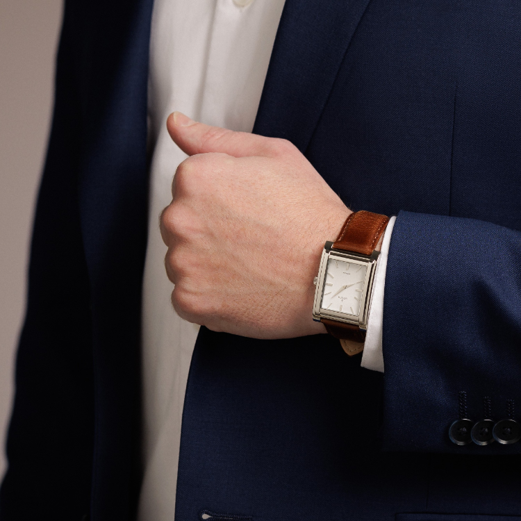 A person in a blue suit jacket and white shirt adjusts the strap of a rectangular watch, featuring a brown leather band. Only the forearm is visible.