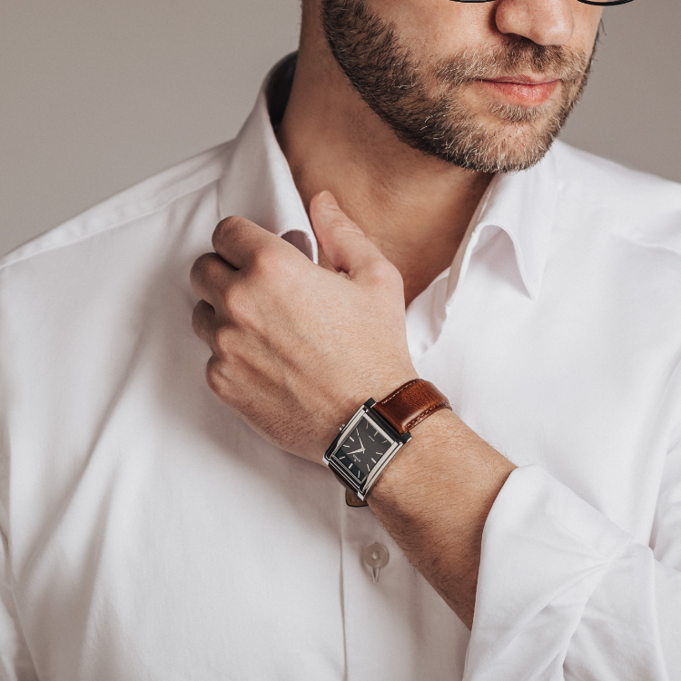 Man in a white dress shirt adjusts his collar with one hand. He has a neatly trimmed beard and wears glasses. On his wrist is a watch with a black square face and brown leather strap. Background is a simple gray.