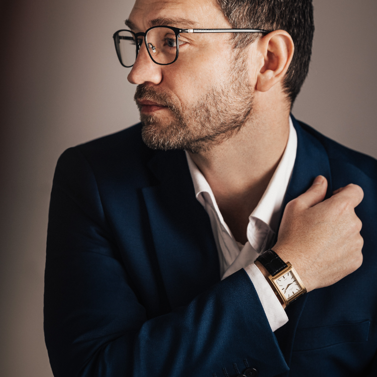 A man with a beard and glasses wearing a blue suit and white shirt looks to the side. He holds his arm, displaying a rectangular watch with a black band. The background is a gradient of dark and light tones.