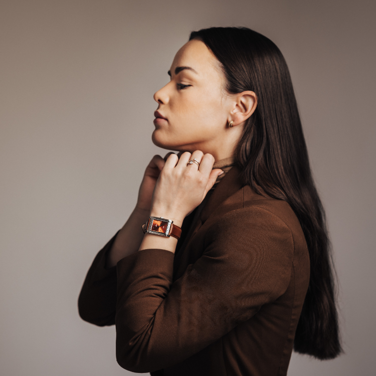 A woman with long dark hair adjusts a necklace. She is wearing a brown blazer and a rectangular watch with a red face. The background is a neutral tone.