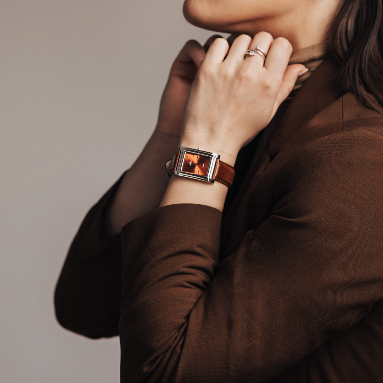 A person in a brown blazer adjusts their turtleneck, showcasing a rectangular watch with an orange face and a leather strap. Only part of the face is visible, drawing attention to the watch and the clasped hands.