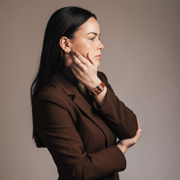 A person with long dark hair and a brown outfit is seen in profile against a neutral background, one hand on their cheek and the other crossed across their body, showcasing a sleek rectangular watch on the left wrist.