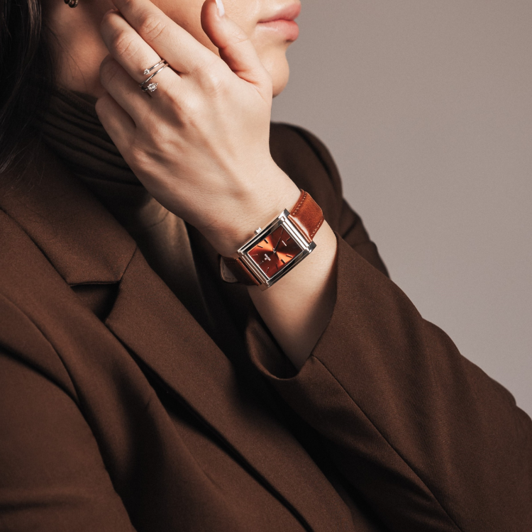 A person in a brown blazer and turtleneck touches their face, elegantly showcasing a rectangular watch with a brown leather strap. A ring glints on their hand against the neutral background.