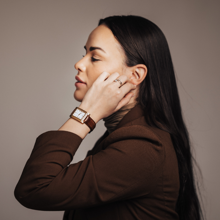 Woman with long dark hair in a brown blazer touching her ear, wearing a rectangular brown leather watch and rings, against a neutral background.