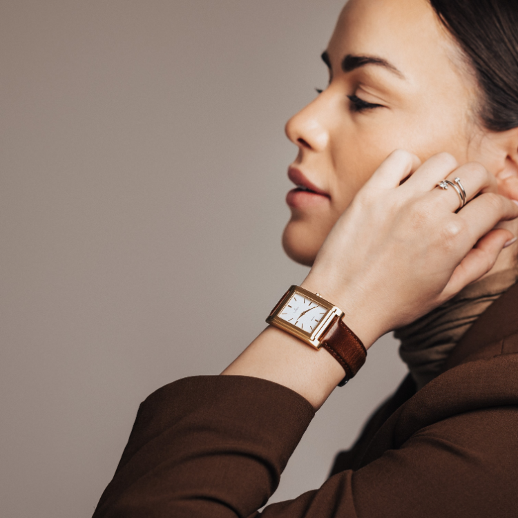 Person with dark hair is shown in profile, eyes closed, wearing a brown coat. Their hand touches their neck, displaying a gold wristwatch with a brown leather strap. The background is plain and neutral.