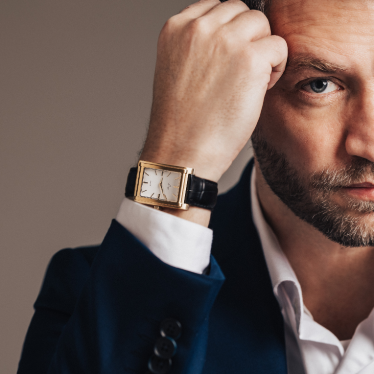 Man in a dark suit with a white shirt, partially visible, resting his head on his hand. Hes wearing a rectangular gold watch with a black leather strap on his wrist, in a neutral, softly lit background.