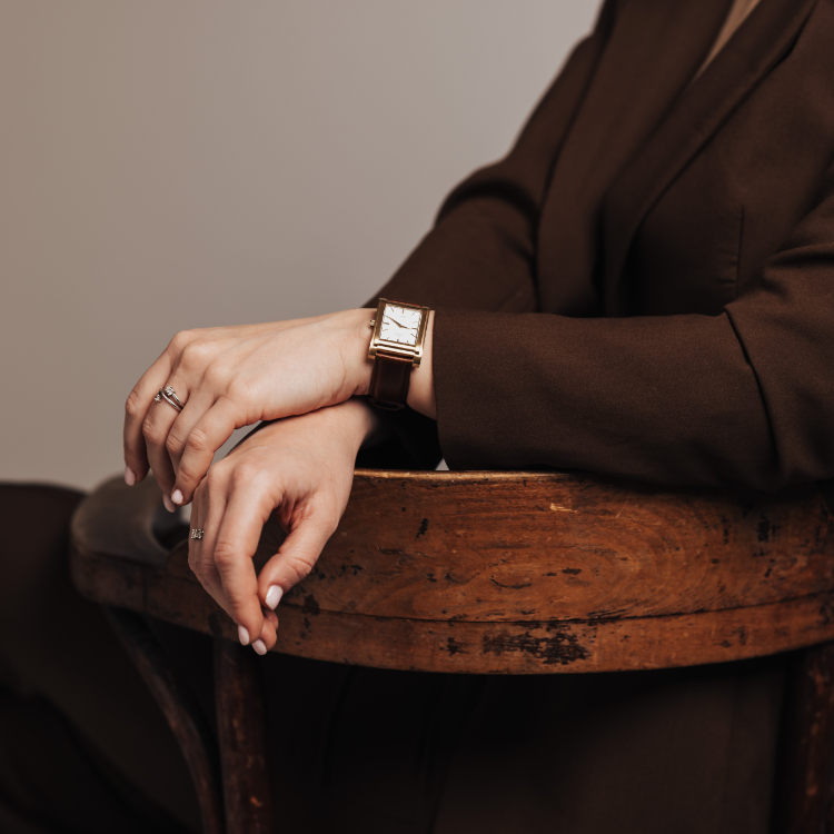 A person in a brown suit sits on a wooden chair with arms crossed, showcasing a sleek rectangular watch. Their hands rest gently, adorned with rings on both index fingers. The focus is on the unique watch and the elegant positioning of the person’s hands.