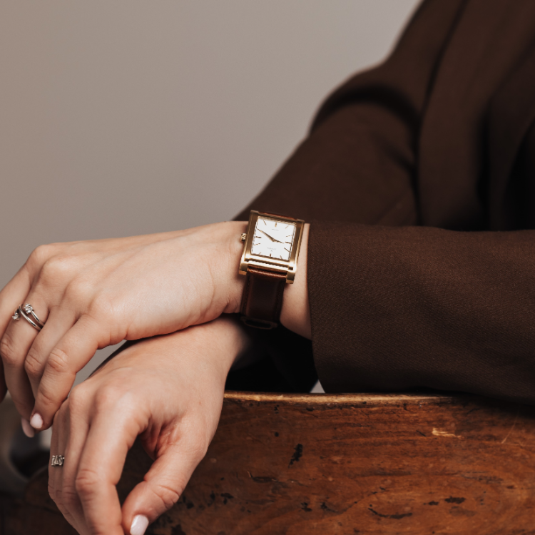 A person wearing a brown jacket rests their hands on a wooden chair. They have a ring on one hand and a rectangular watch with a brown leather strap on the wrist. The background is plain and softly lit.