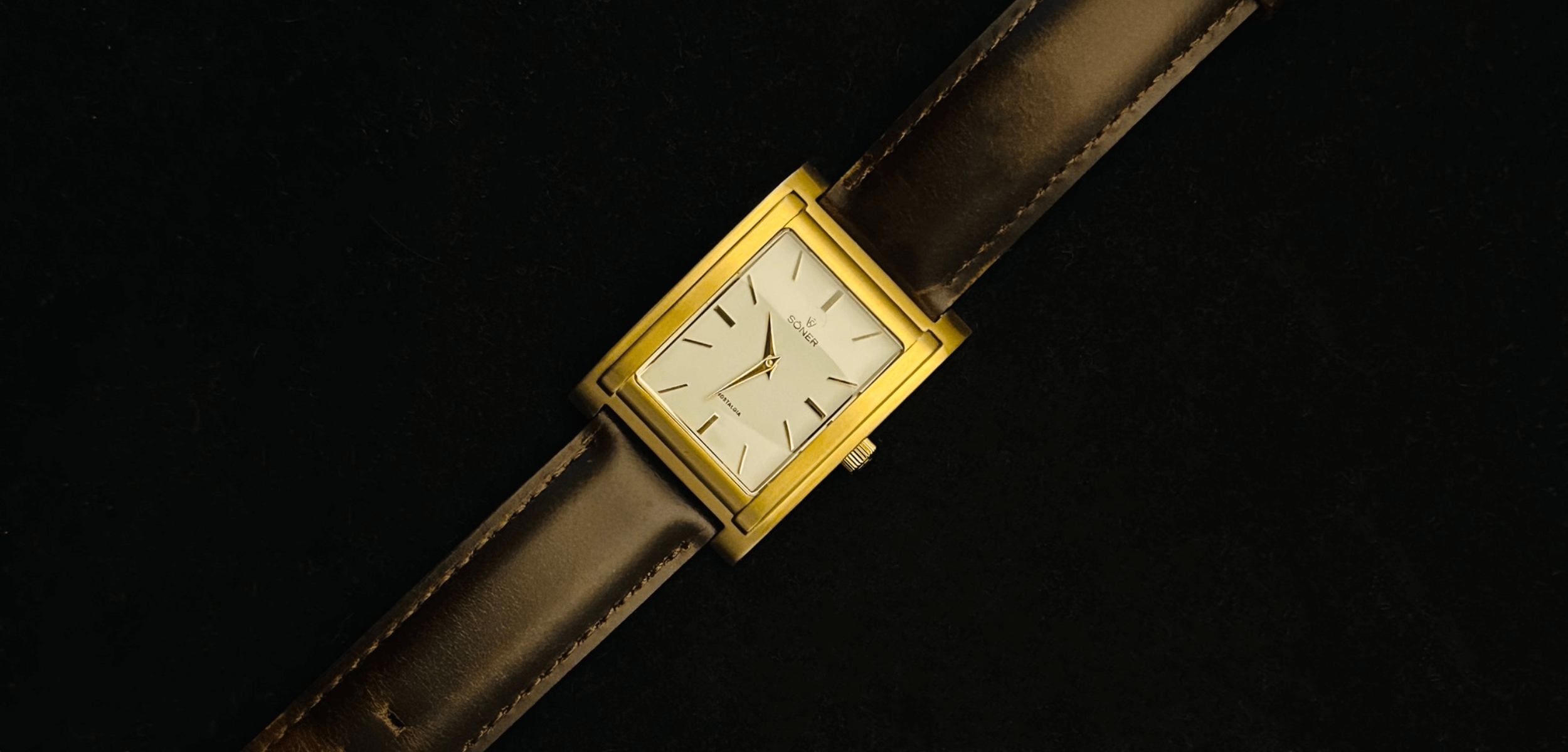 A rectangular gold wristwatch with a white face and minimalist black hour markers is set against a dark background. It has a brown leather strap. The watch hands indicate the time is just past two oclock.