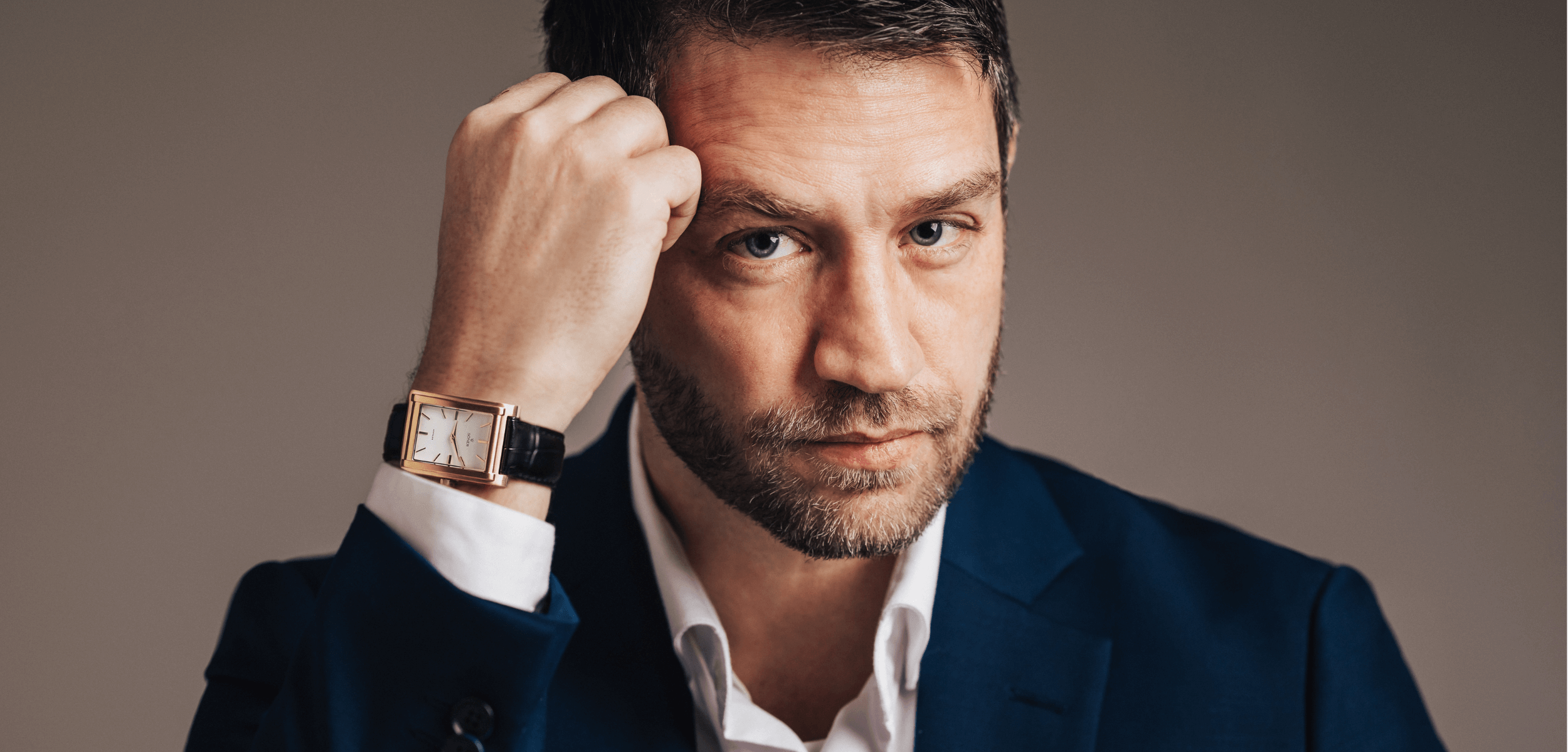 Man with short hair and a beard dressed in a blue suit and white shirt, showcasing a sleek rectangular watch with a leather strap on his raised wrist, gazes intently at the camera against a neutral backdrop.