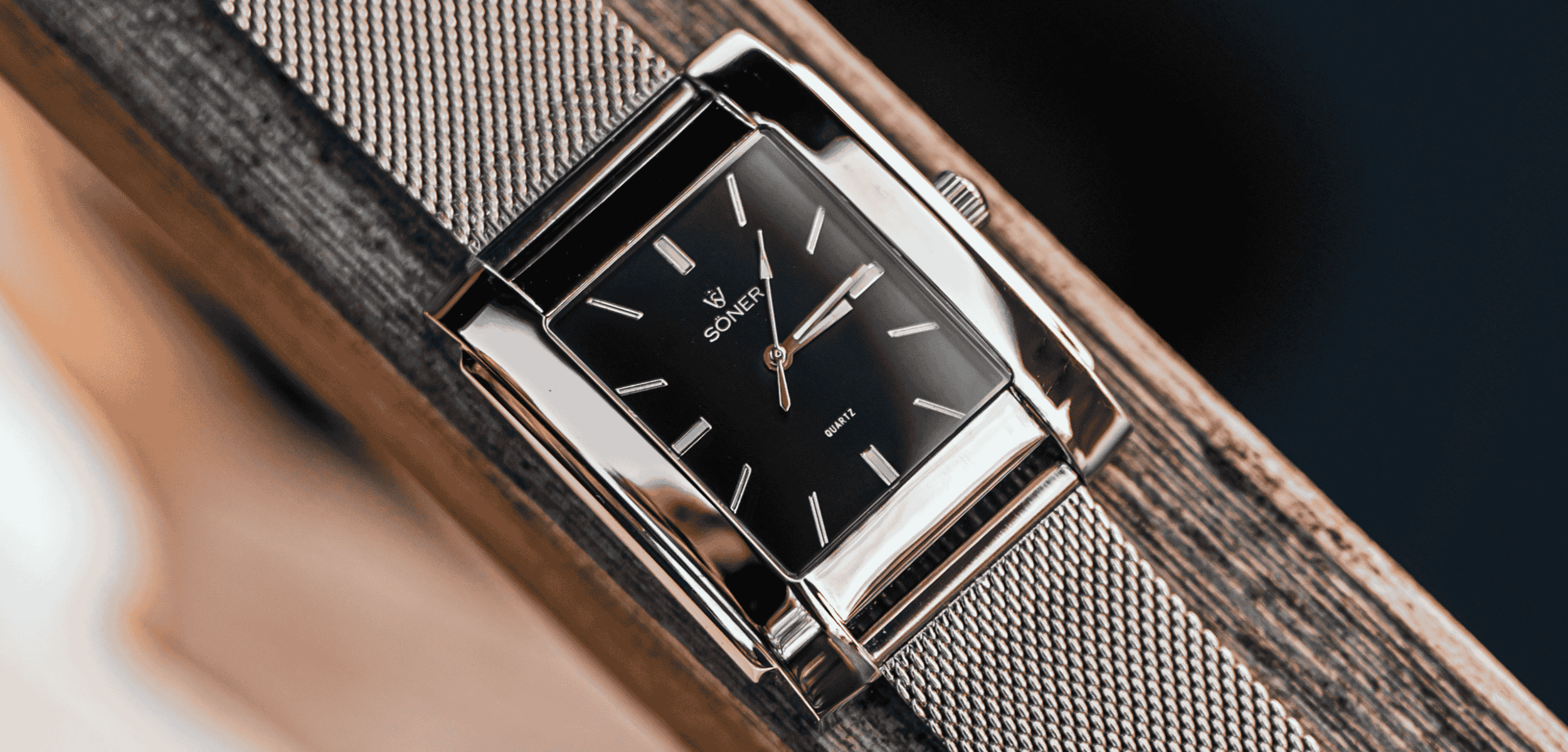 A square black dial wristwatch with silver hour markers and hands, enclosed in a silver rectangular case. The timepiece has a mesh metal strap and rests on a wooden surface, angled to highlight its sleek, modern design.